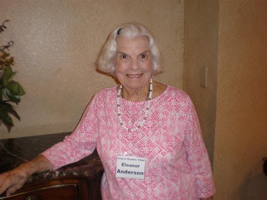 An elderly woman in a pink dress smiles, showing off her handmade necklace at a community event.