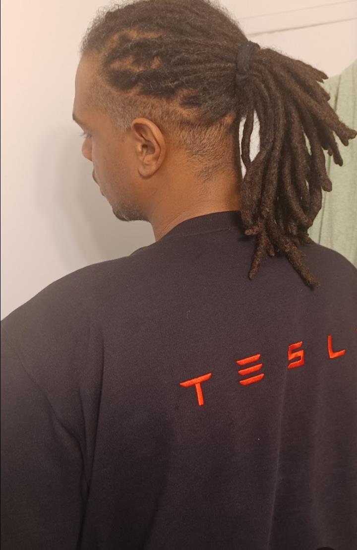 A man with dreadlocks is seen from behind, wearing a shirt featuring the word T E S L.