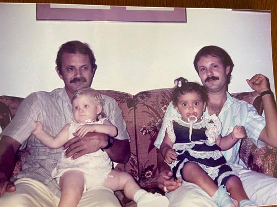 Two fathers sit on a couch holding their young children, enjoying a family moment together.