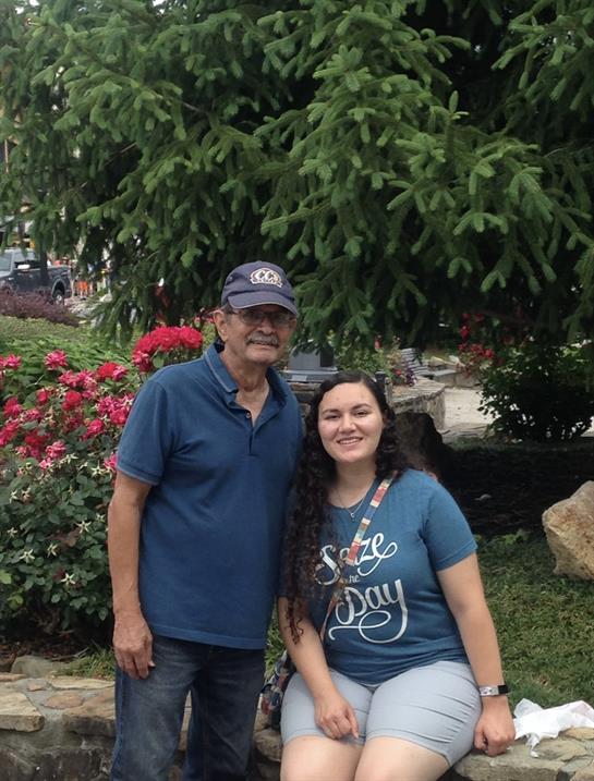 Two people stand together, smiling joyfully in front of vibrant flowers on a sunny day.