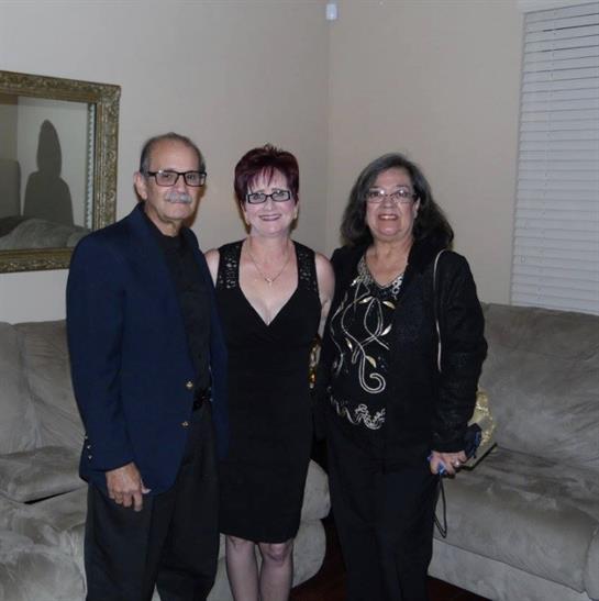 Three adults stand together in a warm living room, dressed in elegant attire for a celebration.