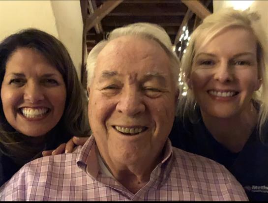 Two women smile warmly beside an older man, celebrating their family bond in a cozy indoor space.