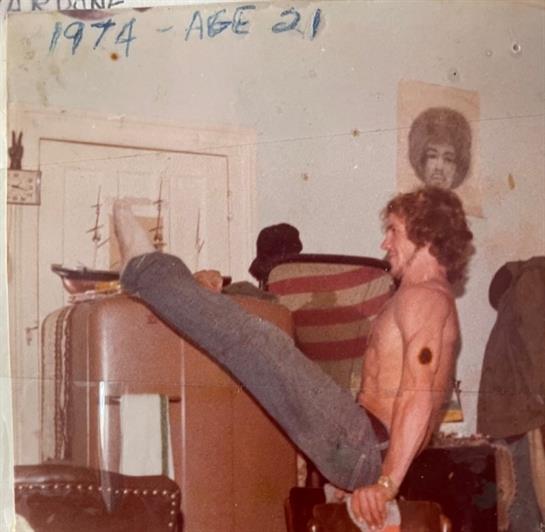 A shirtless young man is exercising with his legs elevated in a retro living room from the 1970s.