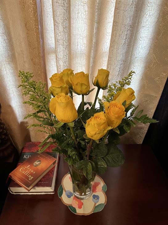 Bright yellow roses arranged in a vase, placed on a wooden table next to soft curtains.
