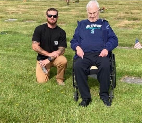 Two men share memories in a cemetery, one in a wheelchair and the other kneeling beside him.