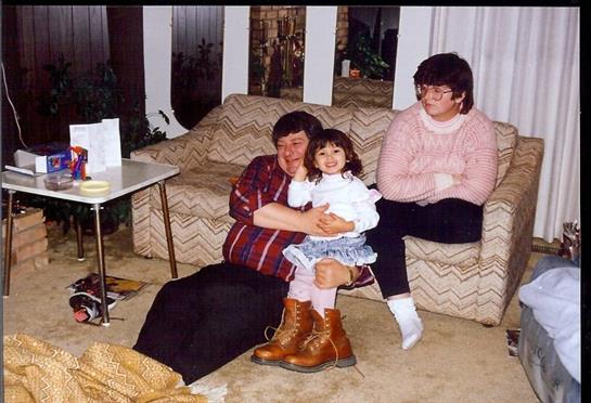 A girl smiles on an adult's lap as another adult watches in a warm setting.
