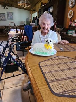 Elderly woman smiles happily beside a decorated cake while seated at a dining table.