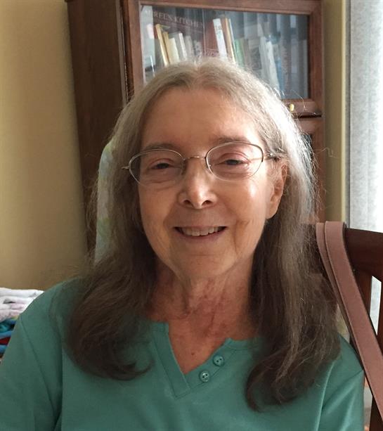 A cheerful elderly woman sits comfortably in her chair, smiling warmly in a bright room.