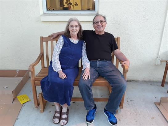 Two adults smile together on a wooden bench in a peaceful outdoor setting.