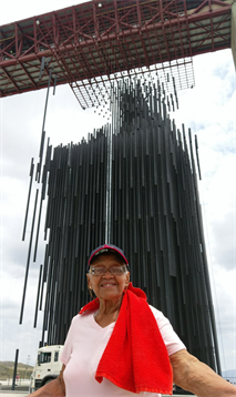 An elderly woman with a red scarf smiles while standing before a large contemporary sculpture.