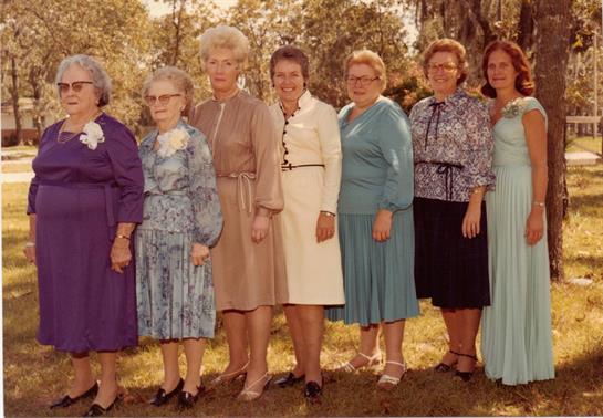 Group of women wearing distinctive dresses smile and pose together outdoors during daylight.
