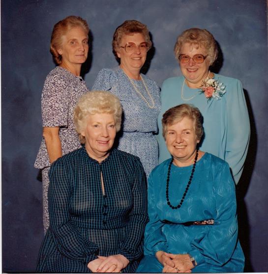 Five women smile together in a formal setting, wearing colorful dresses, enjoying the moment.