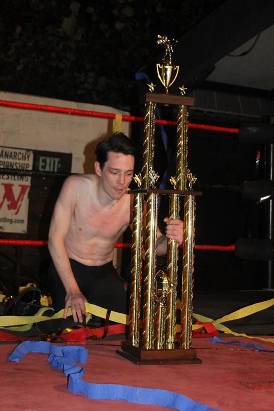 A happy wrestler kneels by a tall trophy, surrounded by ribbons after winning a competition.