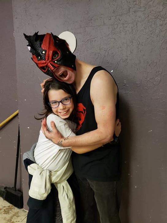 A child joyfully hugs a performer wearing a colorful mask in a community center setting.