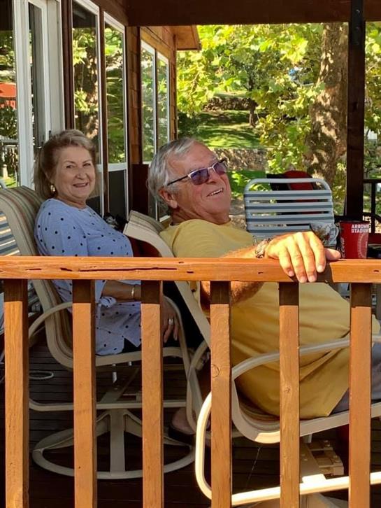 Two people sit on a porch, smiling and enjoying a sunny day while relaxing together.