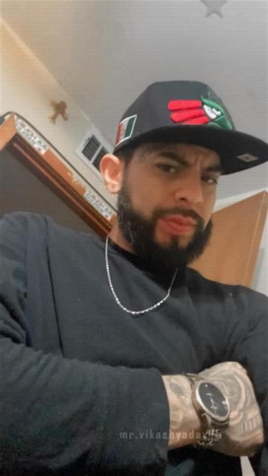 A man stands indoors with a serious expression, wearing a cap and a chain around his neck.