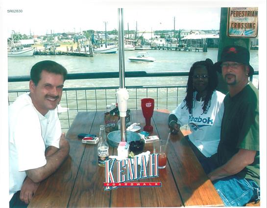 Three men are relaxed at a table with drinks and condiments, enjoying a waterfront view.