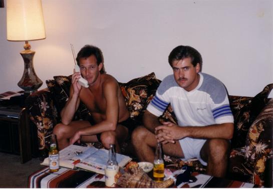 Two men relax on a couch, one speaking on the phone while the other observes, surrounded by snacks.