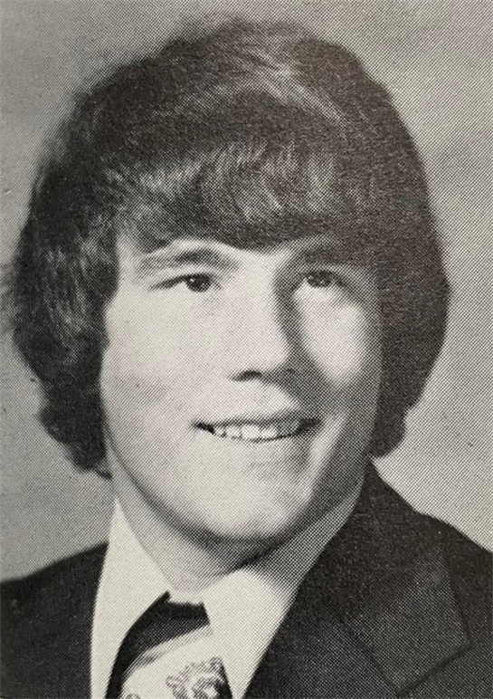 Young man smiles in a senior portrait, with curly hair and formal attire on a neutral backdrop.