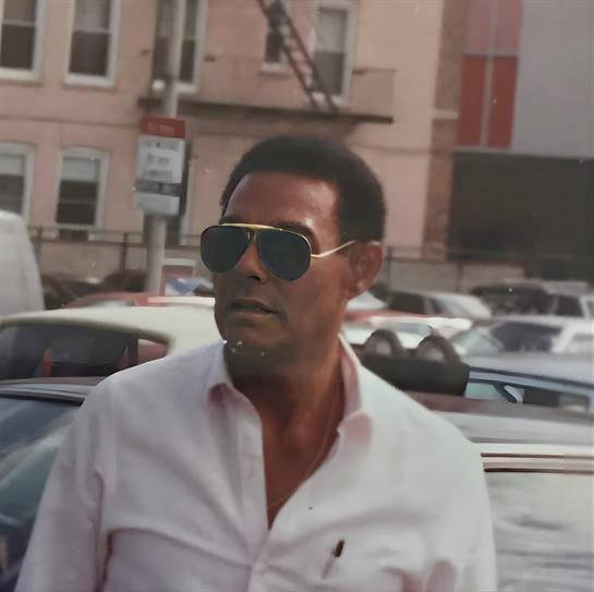 A man in sunglasses strolls past parked cars with buildings in the background.