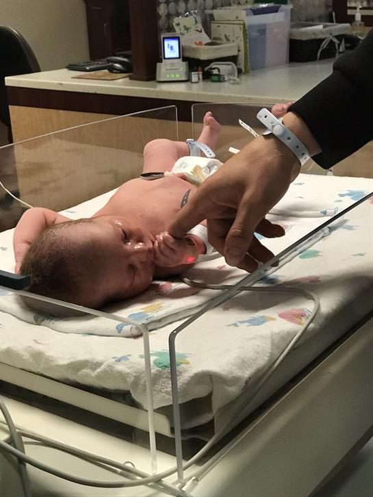A newborn baby lies in a clear bassinet in a hospital nursery, reaching for a caregiver's finger.