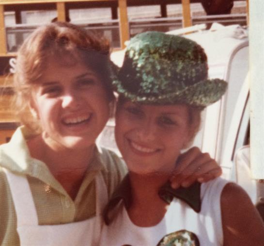 Two young women are joyfully posing for a photo at a fair, wearing vibrant outfits and hats.