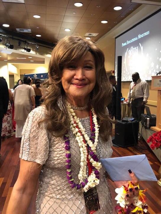 Woman with long hair enjoys a gathering, wearing decorative leis and smiling warmly at attendees.