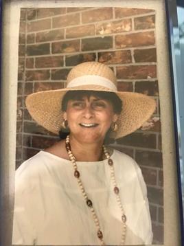 A woman joyfully poses with a wide smile, wearing a straw hat and a necklace near a brick wall.