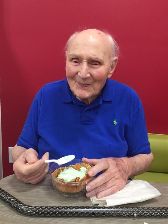 An elderly man enjoys a bowl of dessert while seated at a table in a cafe setting.