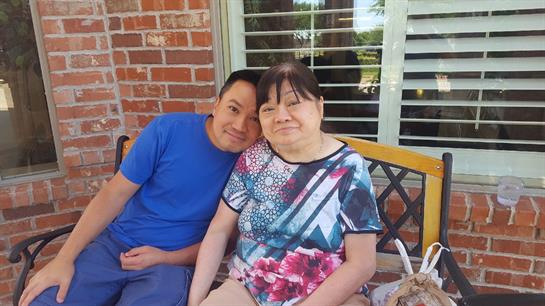 Two individuals relax together on a bench in front of a brick house, sharing smiles.