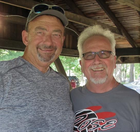 Two men laugh joyfully under a wooden pavilion, enjoying their time at a park event.