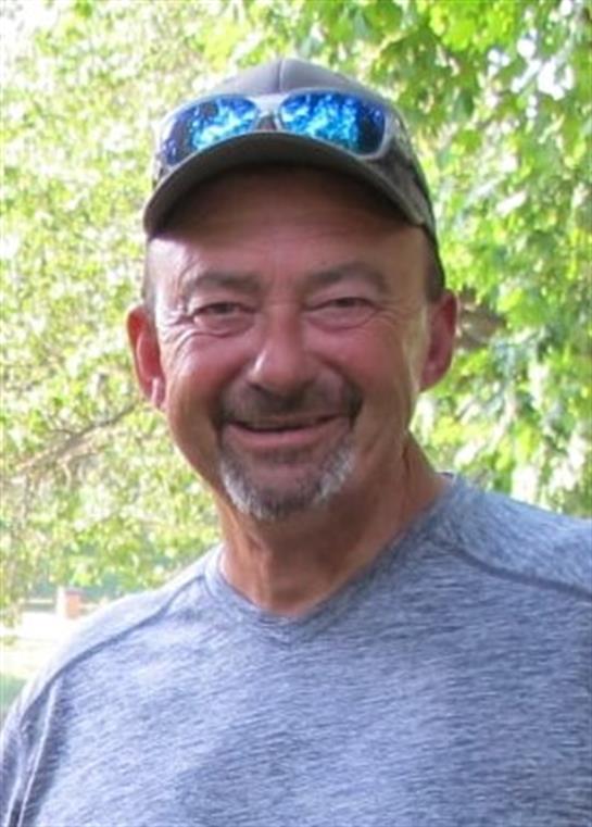 A cheerful man stands outside, surrounded by trees and greenery, enjoying a sunny day.
