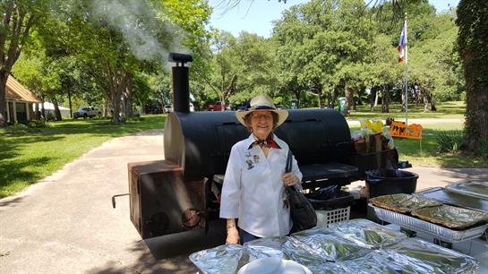 A smiling woman stands by a barbecue smoker in a park, grilling food on a sunny day.