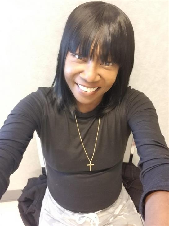 A woman with long hair smiles and poses for a selfie in a simple indoor environment, expressing joy.