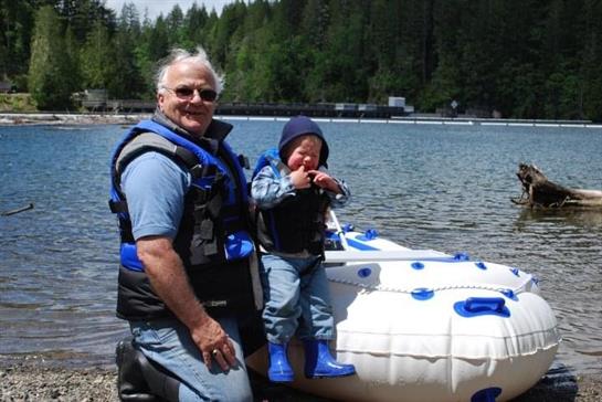A grandfather and his young grandson stand by the lakeshore, both smiling and prepared for kayaking.