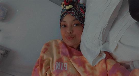 A young woman relaxes on her bed, wearing a colorful blanket and a stylish headwrap while smiling.