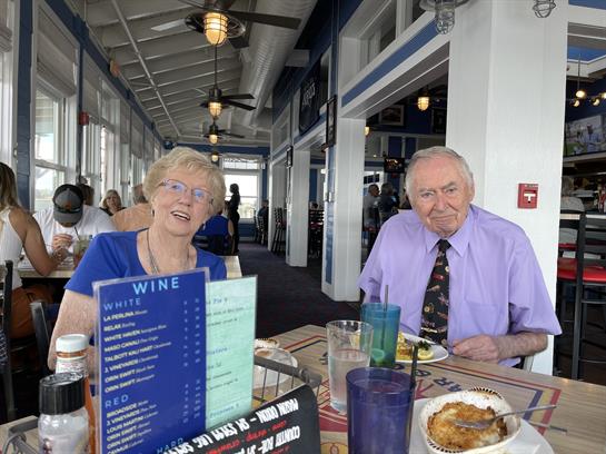 An elderly couple smiles while dining together at a waterfront restaurant, enjoying their meal.