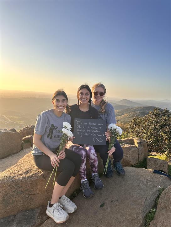 Three friends pose together on a rocky mountain top at sunset, holding flowers and a sign.