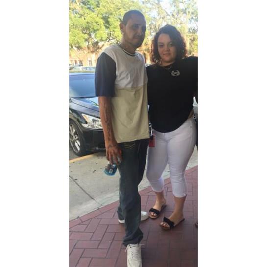 A man and woman stand closely on a sidewalk, smiling and enjoying the moment in bright daylight.