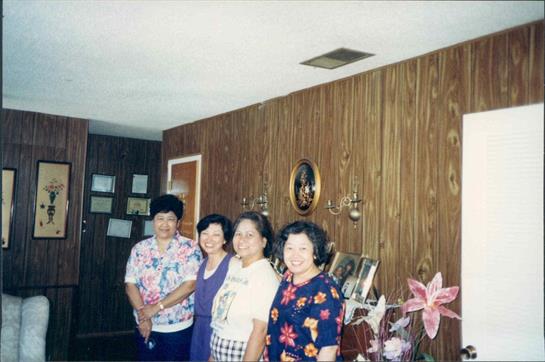 Four women stand together, smiling warmly in a decorated living room, enjoying their time together.