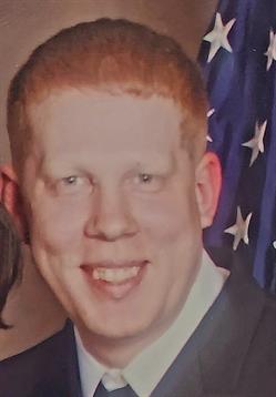 Man with light hair smiles broadly while standing near an American flag at an event.