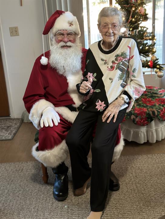 Santa joyfully sits with an elderly woman, surrounded by Christmas decorations and gifts.
