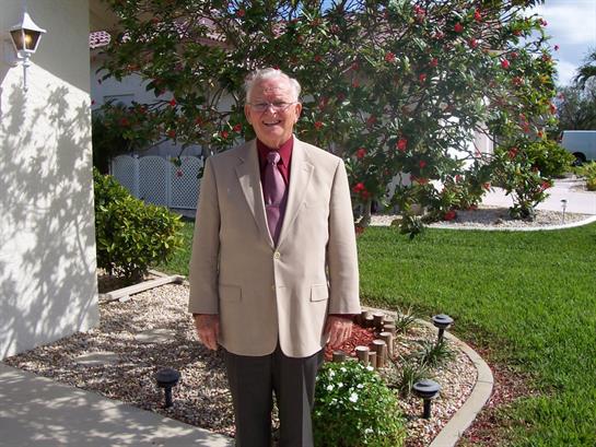 An elderly man wearing a light-colored suit smiles in the garden outside his home.