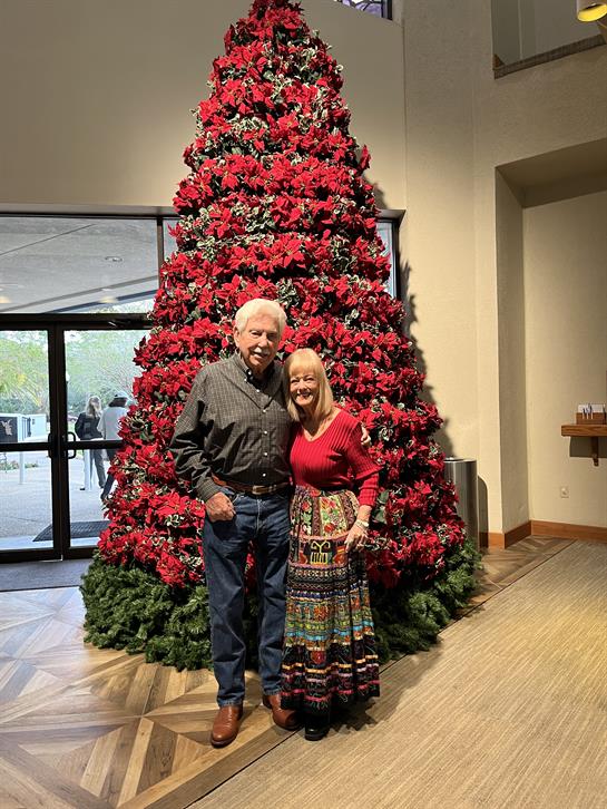 A cheerful couple poses in front of a large poinsettia tree, celebrating the festive atmosphere.