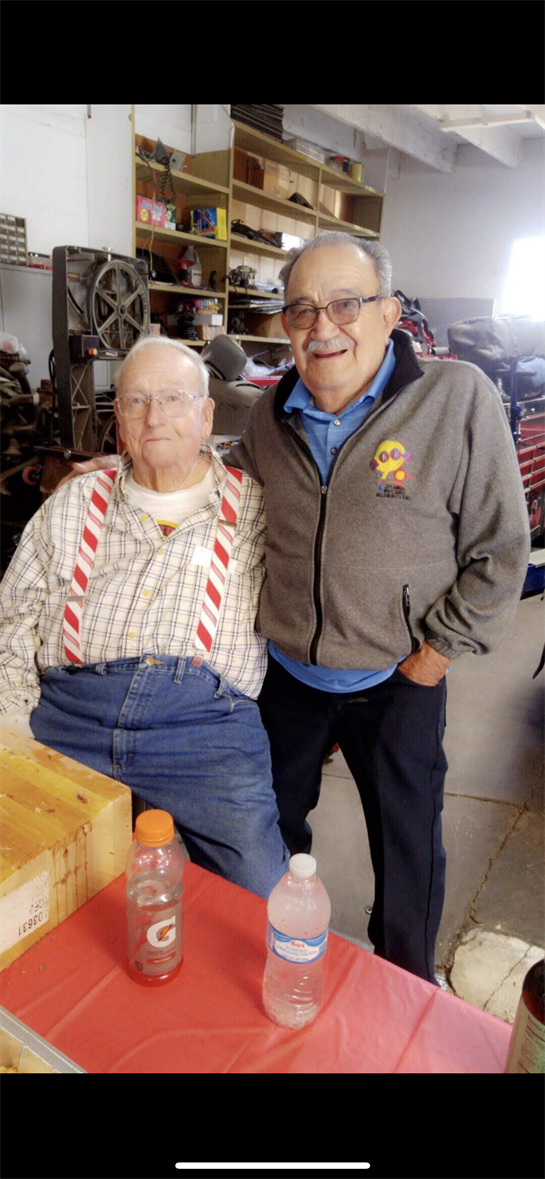 Two older men share smiles in a workshop, surrounded by various tools and equipment.