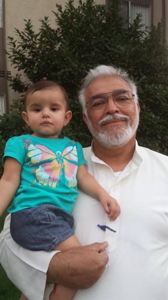A grandfather stands proudly with his granddaughter in a bright green area, both smiling happily.