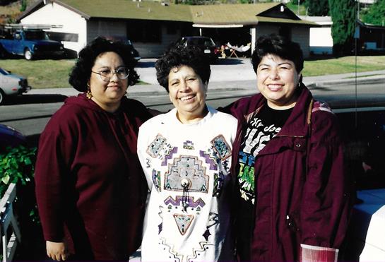 Three women smile and pose outdoors in front of a building on a sunny day.