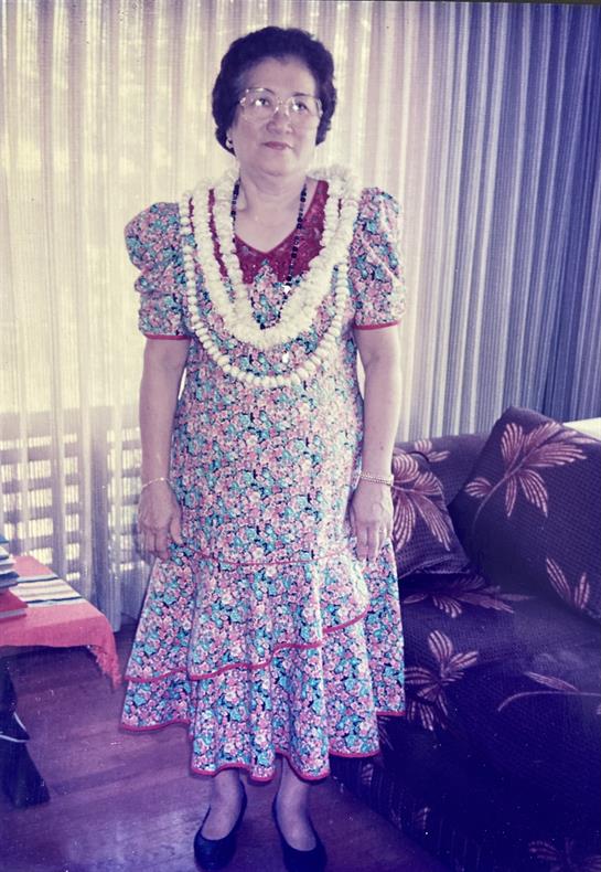 A woman stands in a colorful traditional dress wearing multiple leis in a cozy living room.