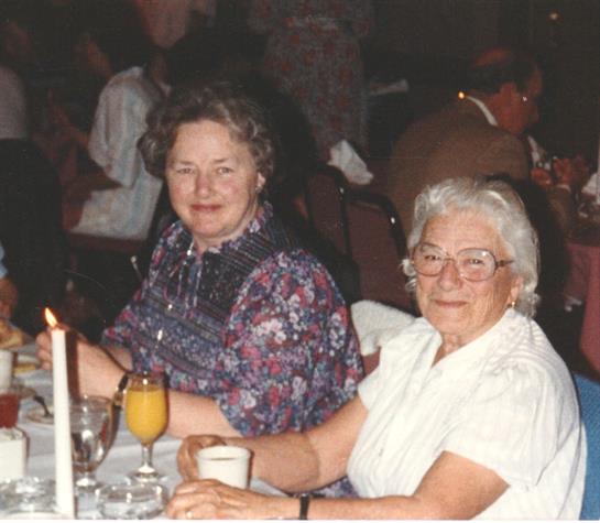 Two seated women smile at the camera, surrounded by a vibrant restaurant atmosphere with dim lights.