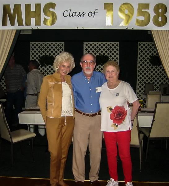 Three former classmates pose together at their 1958 reunion in a festively decorated venue.